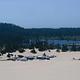 Group of trailers and trucks on a large expanse of sand near a conifer encircled lake under a blue sky.