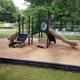 Children's playground with slide, climbing structure, and sandbox, surrounded by a black border and gravel surface, with boats on trailers and lush green trees in the background.
