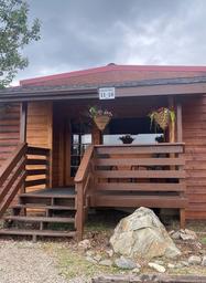 Exterior porch of a wooden cabin with hanging flower baskets and signage for room numbers — clearly shows cabin/motel accommodation on the property (homepage image).