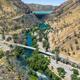 A scenic canyon with a river flowing between steep, rocky hills, a bridge crossing the water, and a winding road lined with trees and greenery.