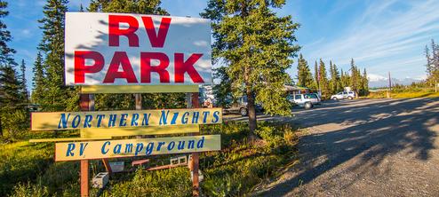 Clear daytime entrance sign reading 'RV PARK / Northern Nights RV Campground' with parked RVs and trees behind — identifies property access and parking area; located on the homepage.