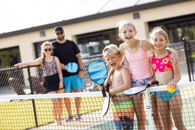 Group of children and adults playing pickleball outdoors, two children holding paddles and smiling, adults standing behind a fence in the background.