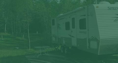 RV parked at a wooded campsite with picnic table and hookups visible — represents the Woods side RV sites and full-service camping described on the site page ('The Woods side').