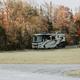 Single RV parked in a grassy site with campground road and tree line in background — illustrates RV pitch size and grounds.