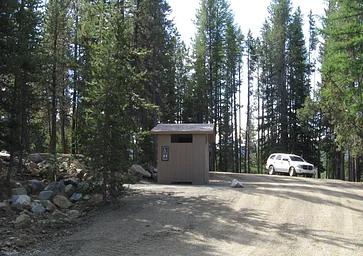 brown vault toilet beside gravel road with white SUV next to pine trees