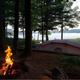 Tent campsite at the water’s edge with a campfire ring and lake visible through trees — illustrates tent camping and waterfront sites (used on Camping Fees page).