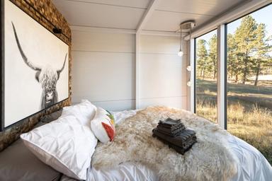 A bedroom with large glass windows showing trees outside. The bed has white bedding, decorative pillows, a furry rug, and a stack of folded dark towels.