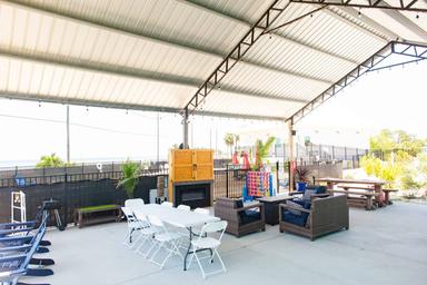 Covered outdoor patio with tables, chairs, sofas, and a fireplace. A large Connect Four game and exercise equipment are visible. String lights hang from the metal roof structure.