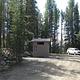 brown vault toilet beside gravel road with white SUV next to pine trees