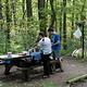 Man and woman standing next to picnic table on wood chip campsite.  On the tent pad behind the campers is a tent.  Waste back hangs from lantern pole.  Site is surrounded by dense forest.