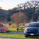 Tent campsite with tent, picnic table and vehicle visible on grassy pad near trees and riverbank — represents tent camping sites and setting on the property.