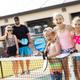 Group of children and adults playing pickleball outdoors, two children holding paddles and smiling, adults standing behind a fence in the background.