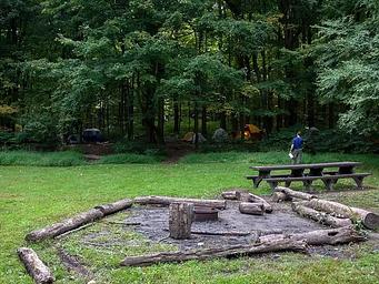 Metal fire ring/grill surrounded by logs and picnic tables in open field.  Multiple tents visible in the distant woods.