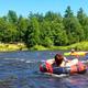 People tubing on a river running through wooded parkland — matches amenities description of river/kayak/tubing (page: https://www.pineridgeparkcampsite.com/amenities). Shows recreational water feature on the property.