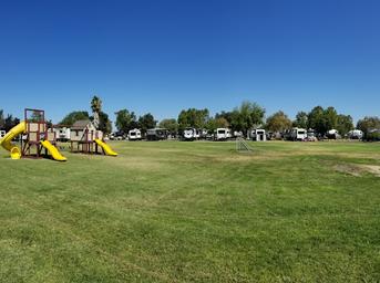 Daytime wide shot showing grassy common area with playground and a row of RVs/cabins in the background — useful overview of campsites and family amenities. Image appears as a background/photo on the site’s home page, indicating it depicts the property.