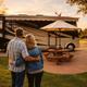 Shows an RV at a paved concrete pad with picnic table and umbrella, and a couple surveying the site — useful depiction of the RV accommodations and site layout.