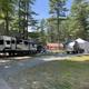 A campground with two RV trailers set up among tall trees. The first trailer is larger with a black and white exterior, an awning, and an American flag. The second is a smaller silver Airstream-style trailer with an attached white canopy. Surrounding