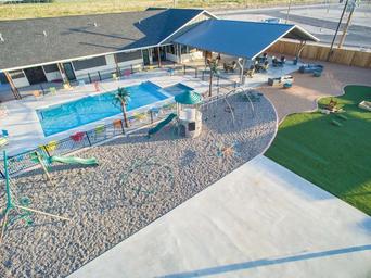Aerial view of a pool area with a swimming pool, a playground with slides and climbing structures, a shaded pavilion with seating, and an adjacent grassy area.