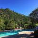 A sunny poolside scene with clear blue water, surrounding trees and hills, lounge chairs, and a bright sky.