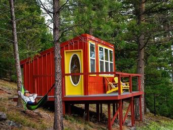 A small, colorful house on stilts in a wooded area, with a red exterior, yellow door, and yellow porch furniture. A person is relaxing in a hammock beside the house.