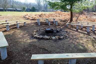 Outdoor fire circle located below athletic field. Metal fire ring/grill surrounded by a stones. Wooden benches on concrete pilings. Rear of dining hall in the background