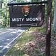 Outdoor wooden sign on road with NPS Arrowhead. Sign labeled Misty Mount with an arrow symbol.