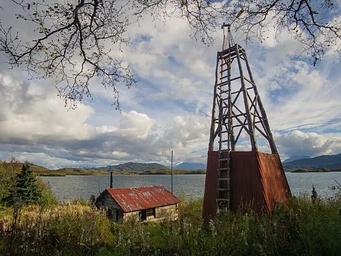 View of a wood cabin and windmill in a grassy field with a lake and mountains in the background.