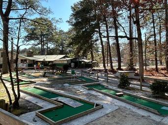 On-ground photo of the mini-golf/course area and clubhouse building near trees and the pond — depicts on-site recreation and facilities.