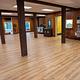 Interior view of dining hall shows 5 large wooden beams, oak laminate flooring, portable a/c unit, pedestal fan, stacked chairs, 6 ft. tables, bottle filling station, waste/recycle bin, and doorway to dishwashing room.