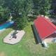 An aerial view of a backyard with a swimming pool, a playground, a red-roofed building, tall trees, and a parking area with RVs and cars.