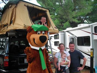 RV campsite area with vehicles and mascot posing — depicts RV parking/pull-through sites and campground setting (homepage gallery image).