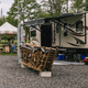 Close view of an RV on a gravel pad with picnic table, firewood stack and utility hookups visible — clearly depicts the RV camping area and site amenities on the property.