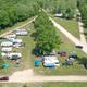 High-resolution aerial overview of the campground showing RV pitches, road layout, central tree row and buildings — matches the site’s ‘campers aerial’ image on the homepage.