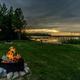 Campfire in a ring on grassy lakeshore with person photographing the sunset — shows campsite atmosphere and firepit amenities by the lake (homepage gallery image).