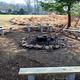 Outdoor fire circle located below athletic field. Metal fire ring/grill surrounded by a stones. Wooden benches on concrete pilings. Rear of dining hall in the background