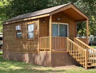 Exterior cabin on-site: clear photo of a wood cabin with porch/steps and neighboring RVs visible, suitable for the cabins listings (appears on the Cabins and Activities pages).