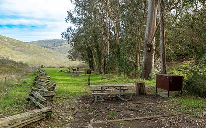 In the foreground are a picnic table and food locker. In the distance are the other campsites, eucalyptus trees, and rolling hills.