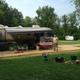 Large RV parked at a long pull-through site with a wooden deck, chairs and fire ring — illustrates the campground’s RV/Park Model accommodations and site setup shown in the ‘ways to stay’ photos.