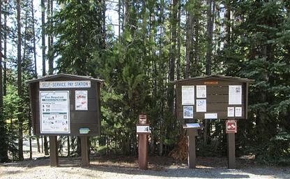 2 information boards with a metal fee vault between and conifers in the background