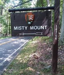 Outdoor wooden sign on road with NPS Arrowhead. Sign labeled Misty Mount with an arrow symbol.
