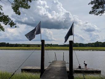 A wooden dock with two flags flying on poles, extending into a Squamscott river. Two people sit and stand on a smaller dock at the end of the main dock, fishing. The sky is partly cloudy with large clouds and neighboring green fields and trees.