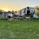 Clear on-site RV campsite view with a fifth-wheel, gazebo, firepit and grassy site at sunset — supports depiction of the campground's RV/seasonal site offerings (from homepage social image).