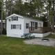 Clear on-site trailer/home with deck, picnic table and fire ring set among trees — matches Camping page context showing rental/park units on Sandy Pines grounds.