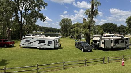 View of RV sites on mowed grass with rigs and vehicles and trees in the background — demonstrates RV camping layout and site spacing.
