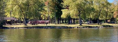 Panoramic shoreline shot showing tree-lined waterfront and campground edge across the water — good aerial/overview-style perspective of the property (used as a background/header on the site).