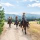 Three horseback riders in western attire ride along a sunny dirt trail lined with trees, open fields, and distant mountain views.
