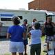 Photo of people gathered next to a parked RV on a gravel site with a brick building in background; appears to show on-site RV campsite/activity and matches the site’s social image (blob-0008.png) on the property pages.