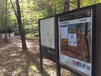 Bulletin board and map sign in the forest near a sidewalk and building in a campground