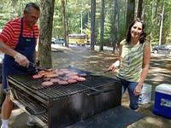 View of two adults cooking on charcoal grill (on site provided) with coolers, parking lot and parked cars in the background.