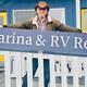 Man in cowboy hat holding a Marina &amp; RV Resort sign outside a blue building.